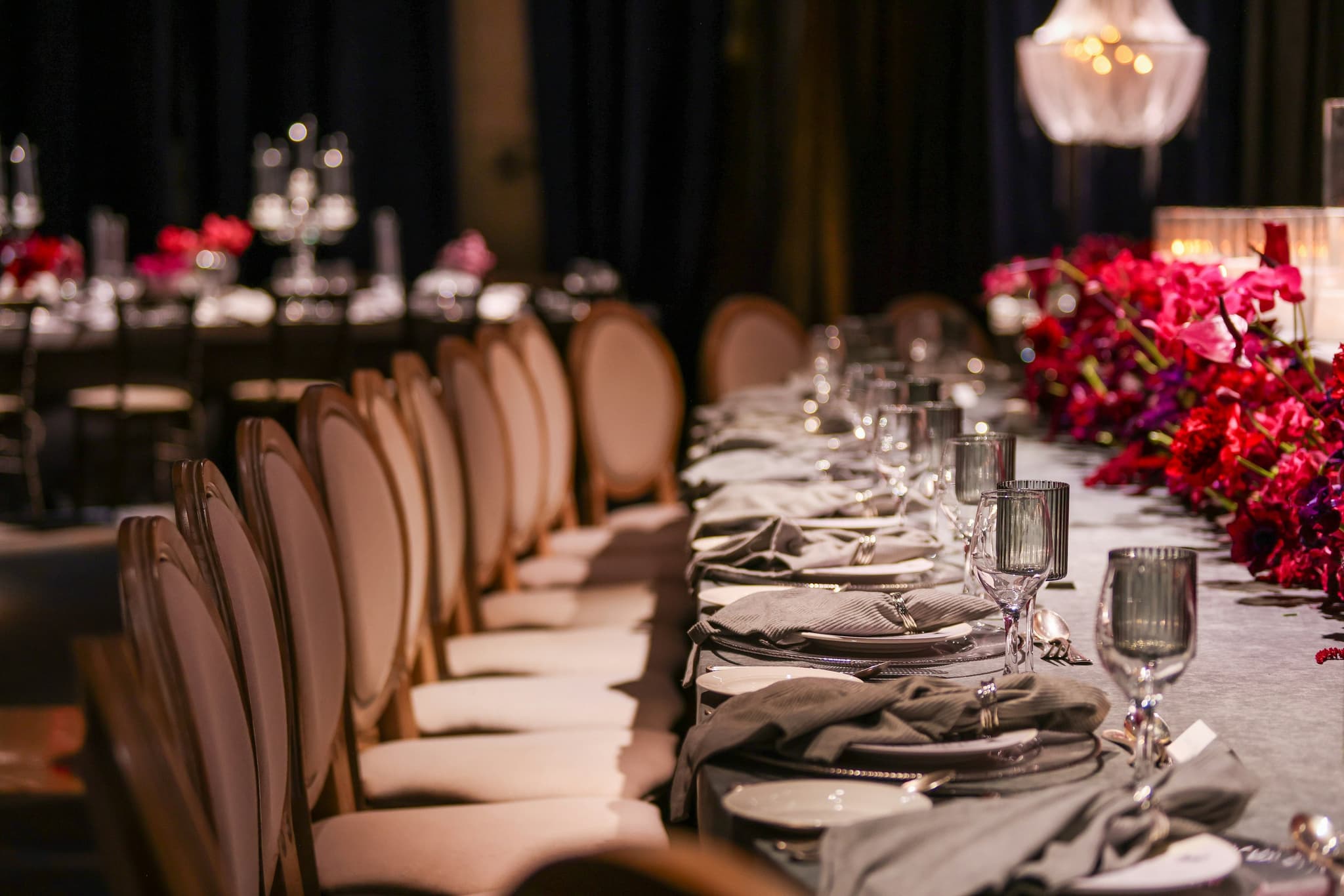 A long banquet table set with glassware and greenery, chairs receding into low light